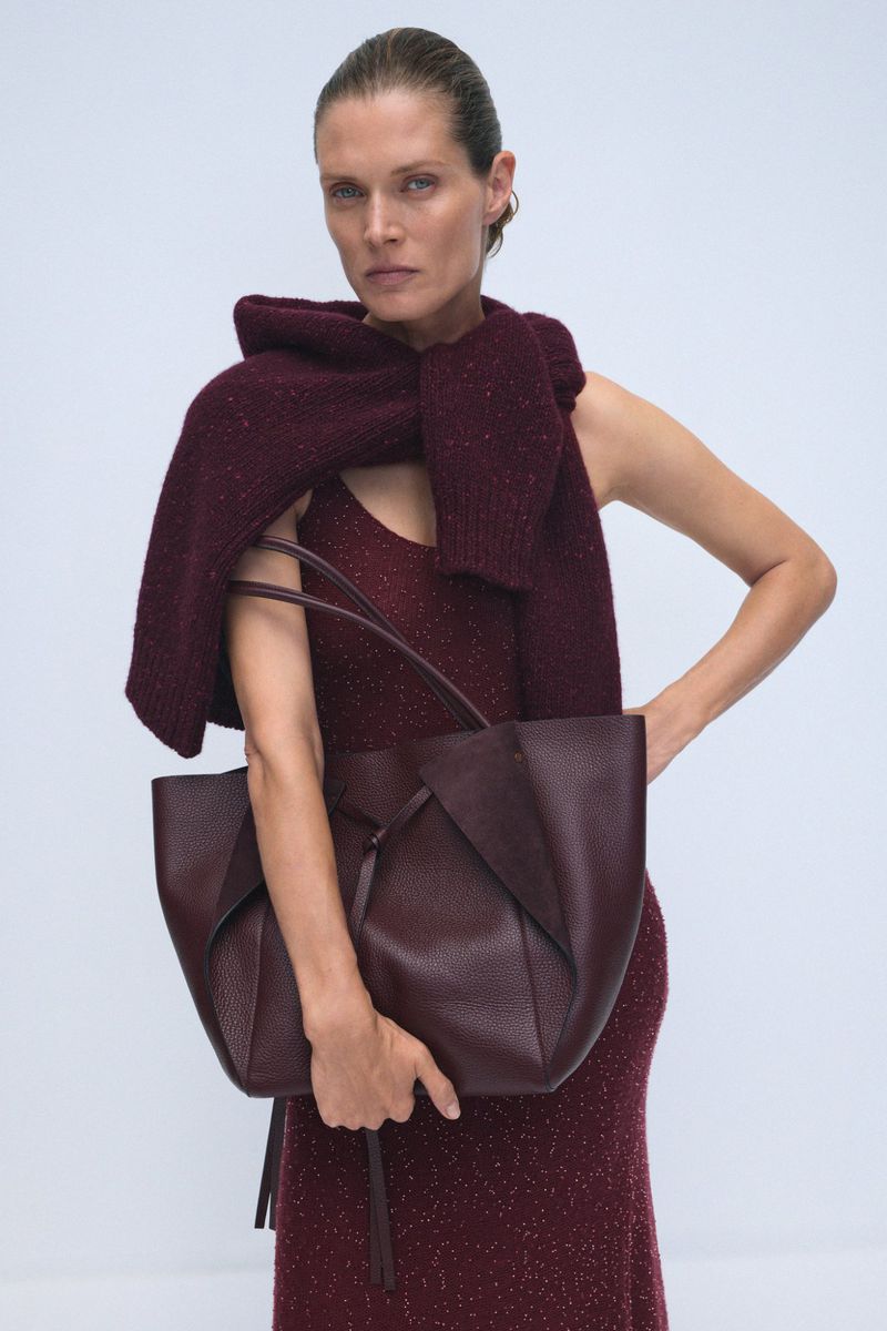A woman models a maroon knit dress with a sweater draped over her shoulders, holding a maroon leather tote bag in a studio shot.