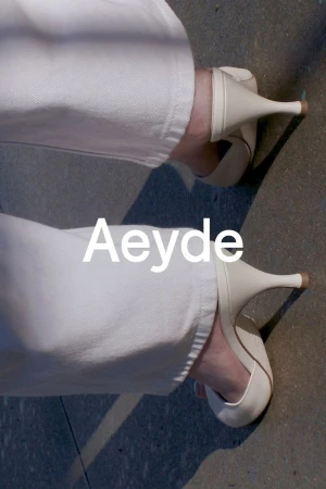 Close-up of woman in white jeans wearing Aeyde brand white kitten heel shoes on concrete surface in studio with light shadows.