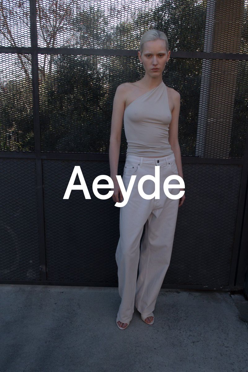 A fair-skinned model poses in front of a dark metal fence, wearing a light tan, one-shoulder top, white pants, and open-toed sandals.