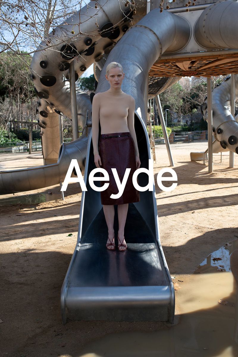 A fair-skinned woman in a burgundy skirt poses on a metal playground slide in a park setting with trees, and the brand name 'Aeyde'.