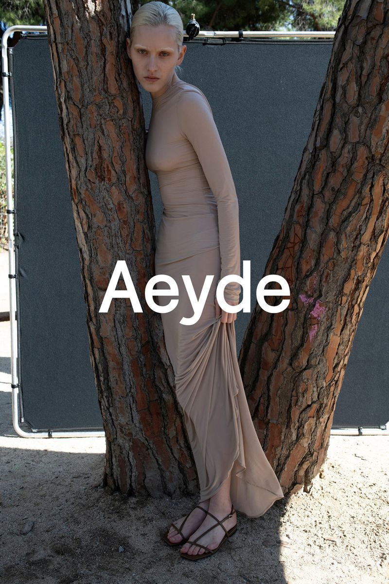 Fashion shot of model posing in a nude dress leaning against a tree outdoors, captured in natural light, with a grey background.