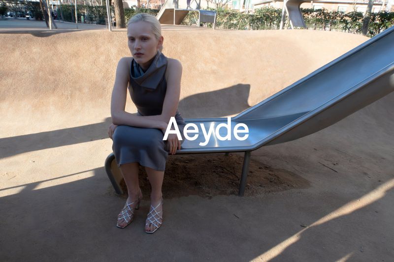 Adult female sitting on a playground slide in a sleeveless grey dress and strappy heeled shoes, outdoor daytime fashion product shot.