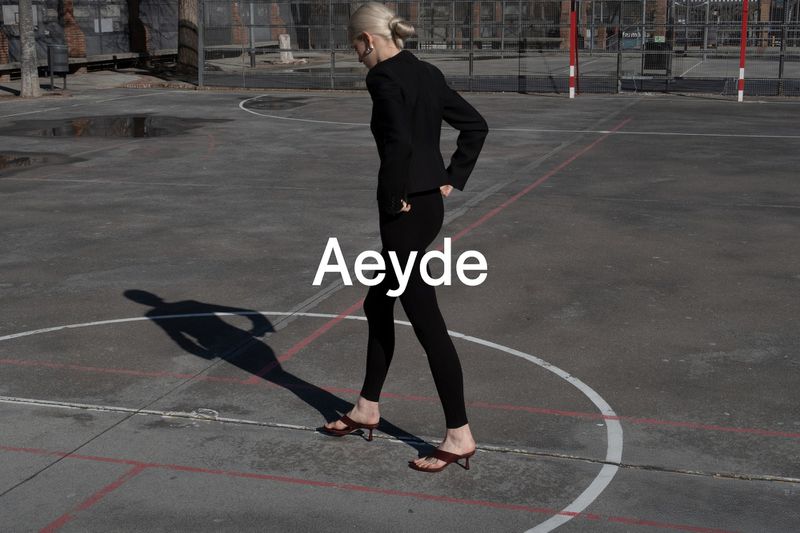 Fashion model in black blazer and leggings walks on basketball court in brown sandals, with Aeyde logo overlayed in white text.