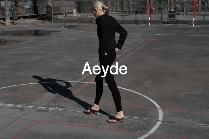 Fashion model in black blazer and leggings walks on basketball court in brown sandals, with Aeyde logo overlayed in white text.