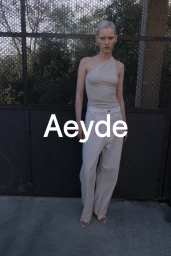 Model wearing a neutral one-shoulder top and light trousers standing against a mesh fence