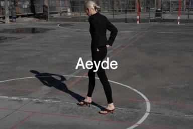 Model in all-black outfit and heeled sandals walking across an outdoor asphalt sports court for Aeyde campaign