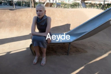 A blonde model seated on a metal slide outdoors wearing a grey dress and strappy sandals for the Aeyde S/S 26 campaign.