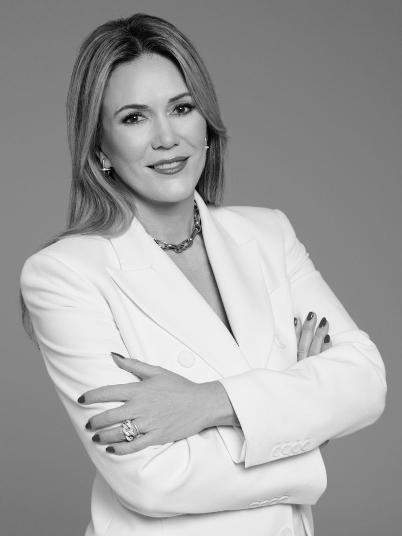 Black and white portrait of a blonde woman wearing a white blazer and necklace with her arms crossed against a grey background.