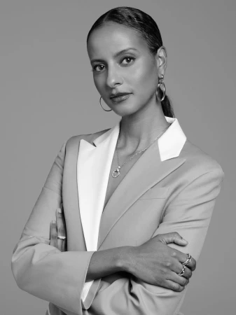 Black and white portrait of an adult female in a blazer with her arms crossed looking at the camera, with gray studio backdrop.