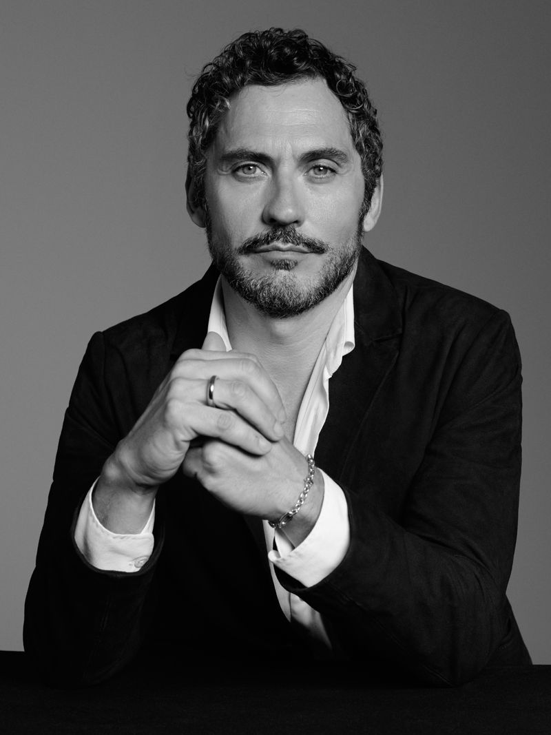 Black and white portrait of an adult male with a mustache and beard, wearing an unbuttoned white shirt and blazer, with clasped hands.