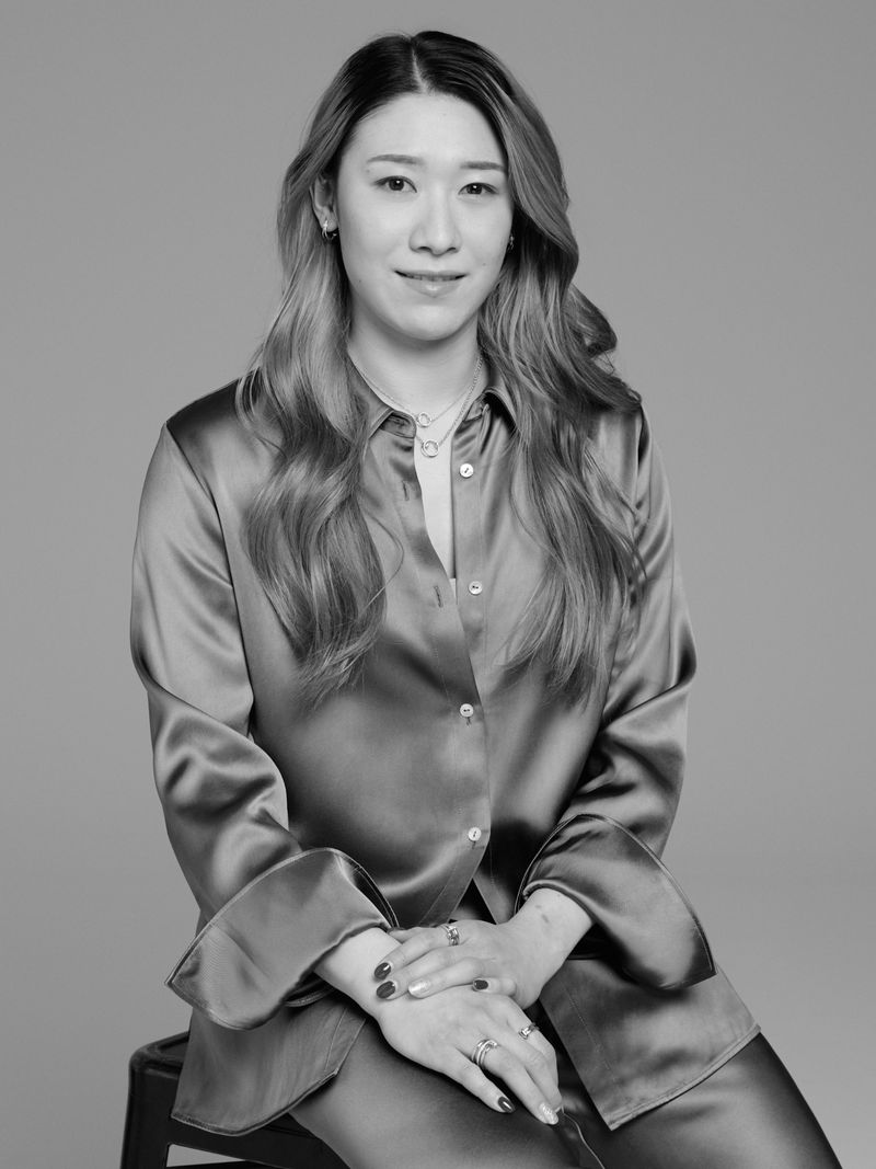 Portrait of an adult woman with wavy brown hair, wearing a satin shirt and pants, seated indoors against a neutral grey backdrop.