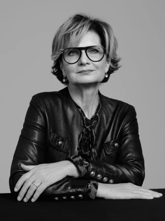 Black and white portrait of a mature woman wearing glasses and a leather jacket, sitting at a table against a neutral background.