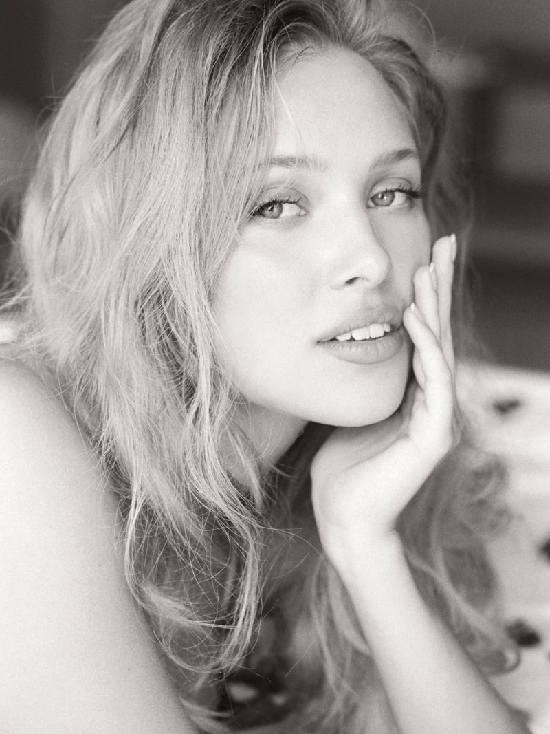 Monochrome portrait of a fair-skinned woman with blonde wavy hair, her hand gently touching her face in a soft, serene pose.