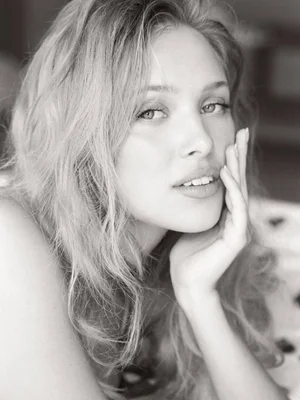 Close-up black and white portrait of a blonde woman resting her face against her hand, looking directly at the camera with a soft expression.