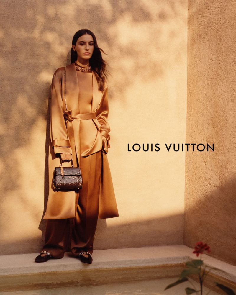 Elegant woman in a bronze satin outfit, accessorized with a necklace and handbag, stands against a textured beige wall with the logo.
