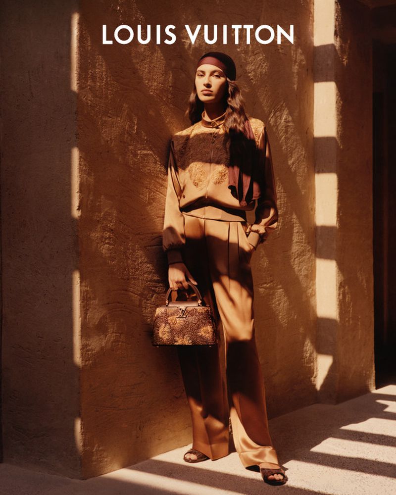 A woman models Louis Vuitton brand tan silk blouse and slacks, holding a structured, printed purse in a studio fashion shot.