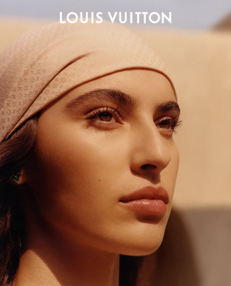 Close-up portrait of a woman with a patterned headscarf looking up, with warm lighting and the Louis Vuitton logo visible.