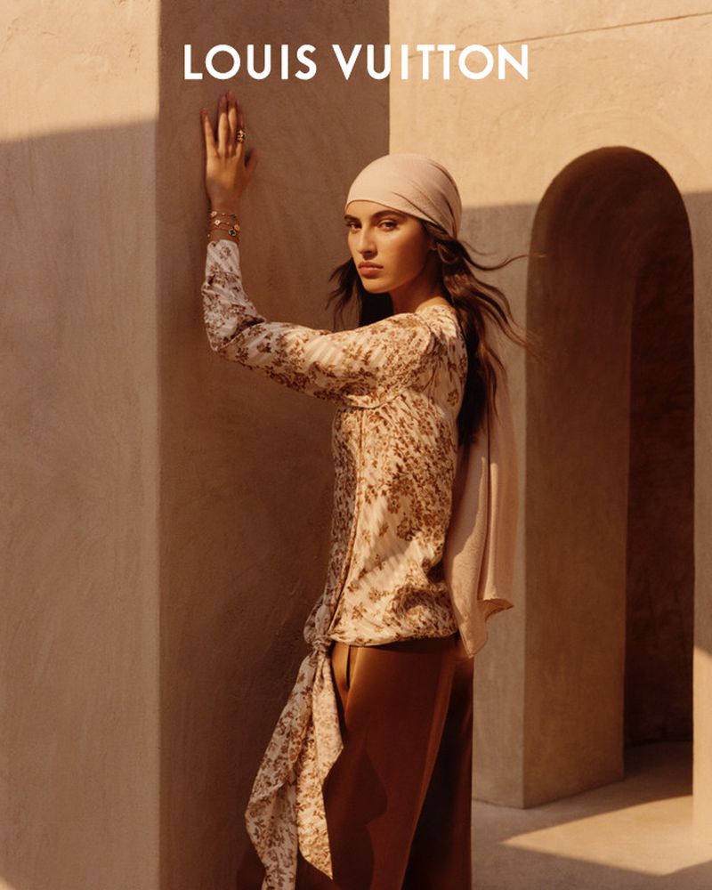 Indoor portrait of a young woman in a patterned top, headscarf, and brown pants, posing in front of a wall with the LOUIS VUITTON logo.