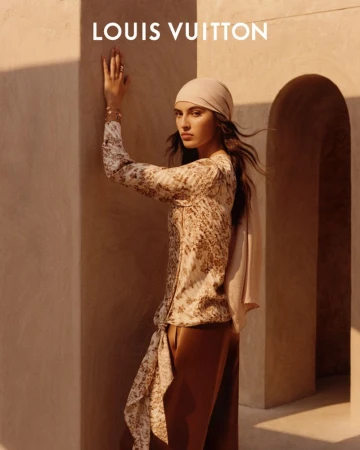 Indoor portrait of a young woman in a patterned top, headscarf, and brown pants, posing in front of a wall with the LOUIS VUITTON logo.