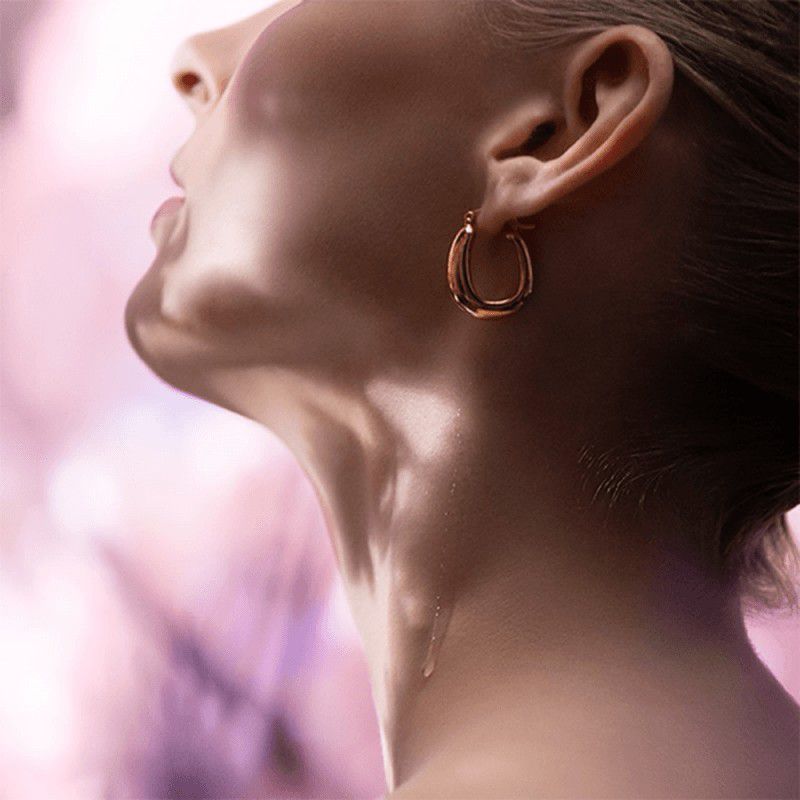 Elegant close-up of a woman's neck and gold hoop earring against a soft, blurred pink background, showcasing beauty and jewelry.