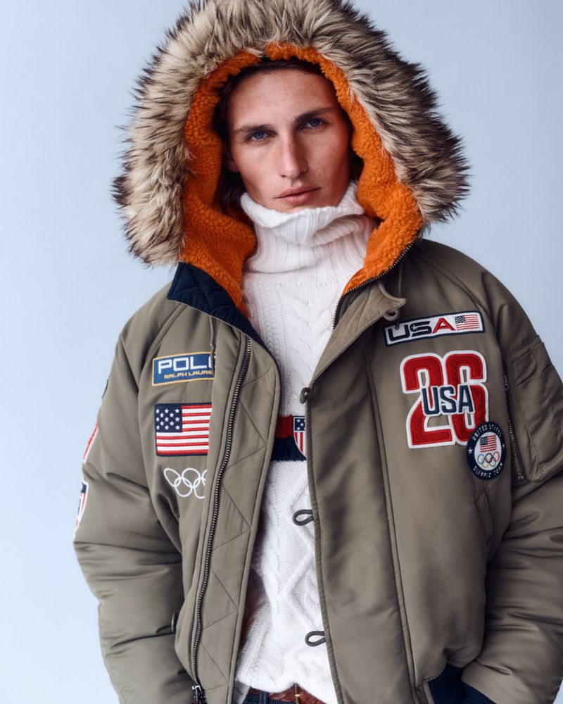 Man in olive green Ralph Lauren puffer jacket with fur-trimmed hood and USA Olympic patches, wearing white turtleneck sweater indoors.