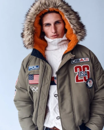 Man in olive green Ralph Lauren puffer jacket with fur-trimmed hood and USA Olympic patches, wearing white turtleneck sweater indoors.