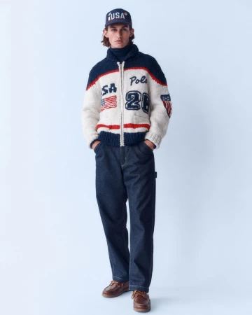 Male fashion model poses in a full shot wearing a navy and white wool zip-up cardigan, jeans, a baseball cap, and brown leather shoes.