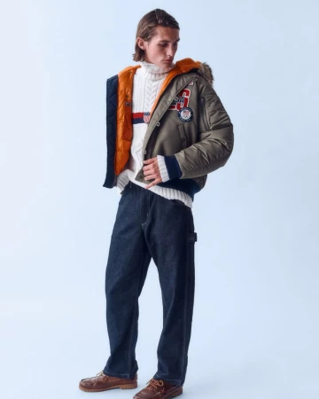 Male model poses in an olive green bomber jacket with a fur-lined hood, paired with a cable-knit sweater, jeans, and leather shoes in a studio shot.