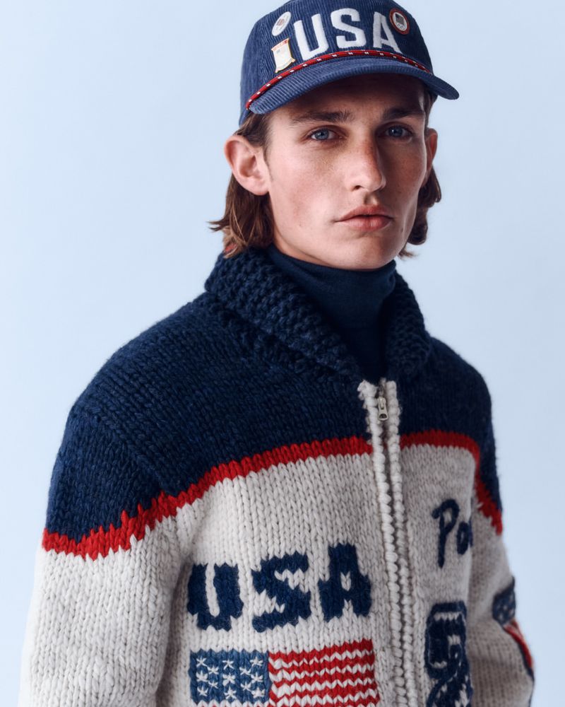 Portrait of a young adult man wearing a baseball cap and a heavy knitted sweater with a USA and American flag motif on a light background.