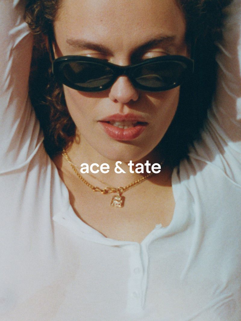 Close-up portrait of a woman wearing dark sunglasses and a gold pendant necklace against a white background