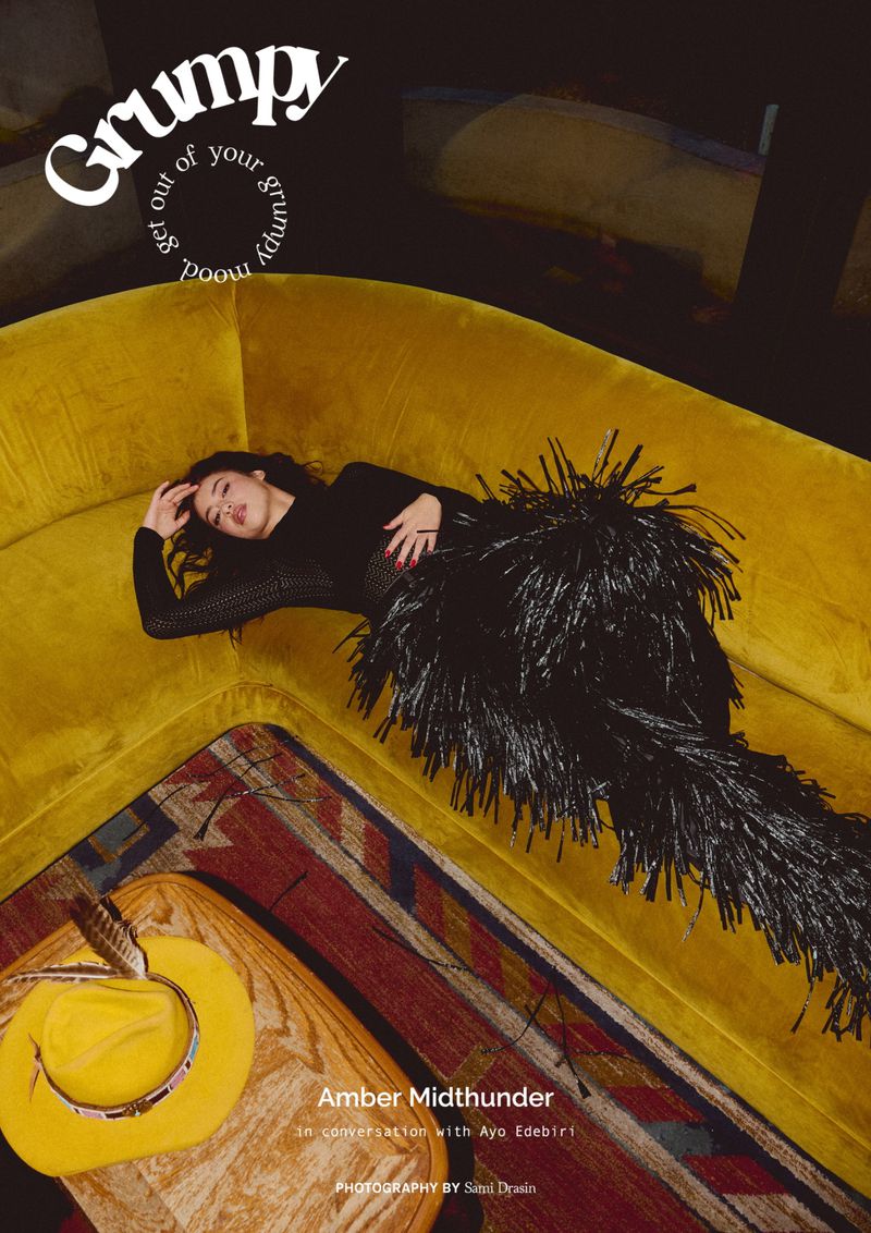 Woman in black dress with feathered skirt reclines on yellow velvet couch with cowboy hat on table in a fashion magazine editorial.