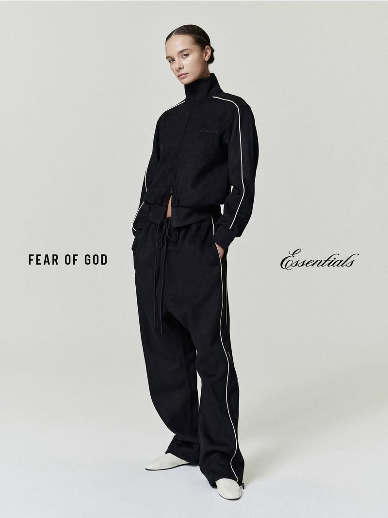 A slender person in a black tracksuit with white piping stands in front of a plain grey studio backdrop, featuring visible brand logos.