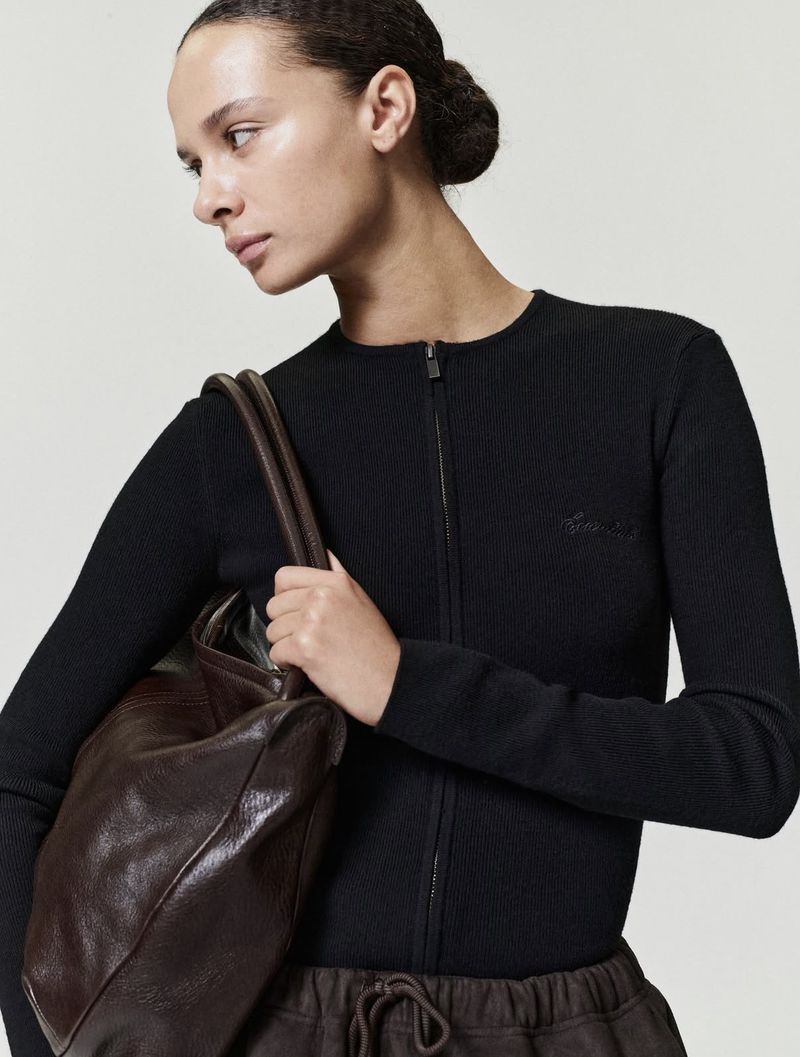 Woman with dark hair holds a dark brown leather bag in front of a plain, light background. She wears a black, long-sleeved, zip-up shirt.