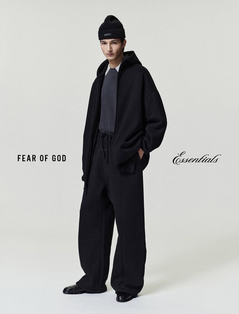 A male model wears a black hoodie, pants, and beanie hat in a full-length studio shot for the brand Fear of God Essentials.