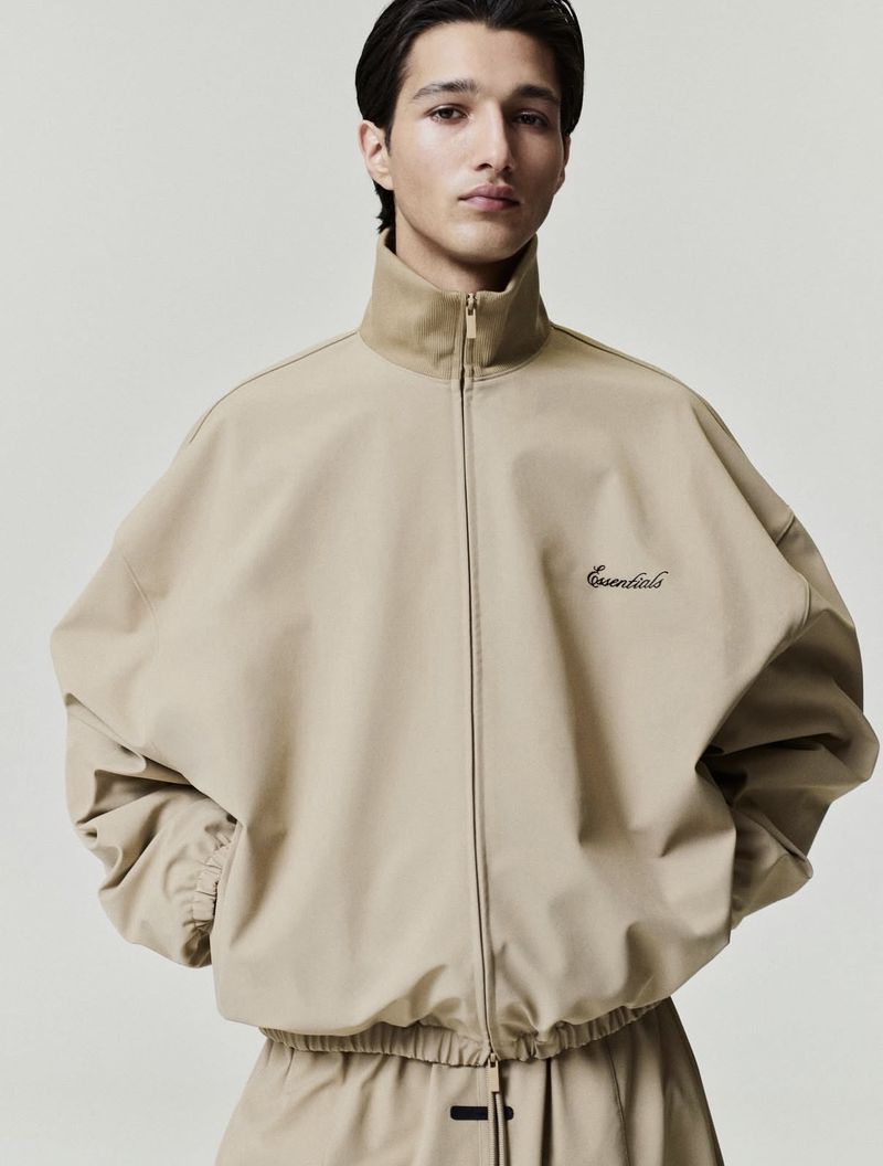 A person poses in a neutral-toned zip-up jacket and matching pants against a plain background, showcasing a relaxed fashion aesthetic.