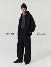 Model wearing a black zip hoodie, matching trousers, and beanie against a neutral light grey backdrop for Fear of God Essentials