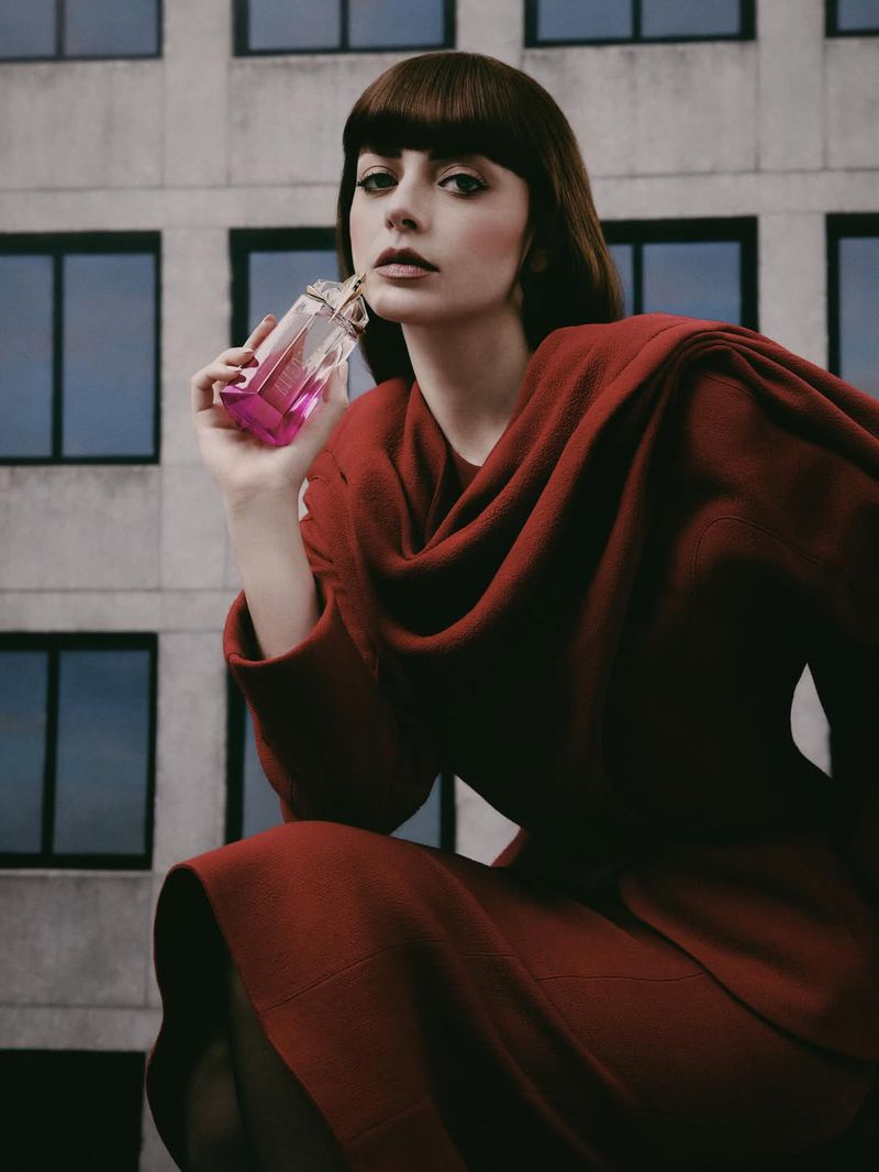 Brunette woman with bangs in red dress holds pink perfume bottle against modern building in this beauty-focused fashion editorial.