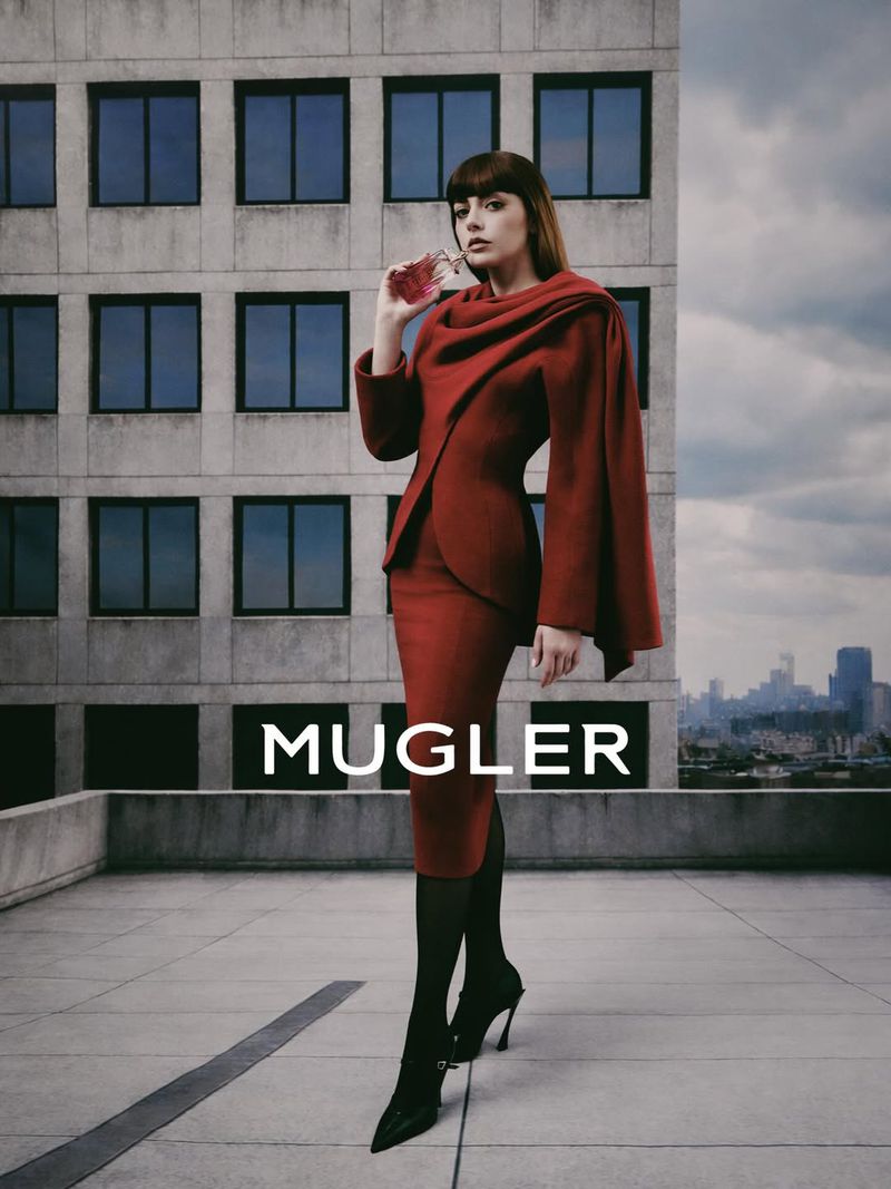 A brunette adult female poses in a red outfit holding a perfume bottle against a city backdrop in a fashion forward portrait.