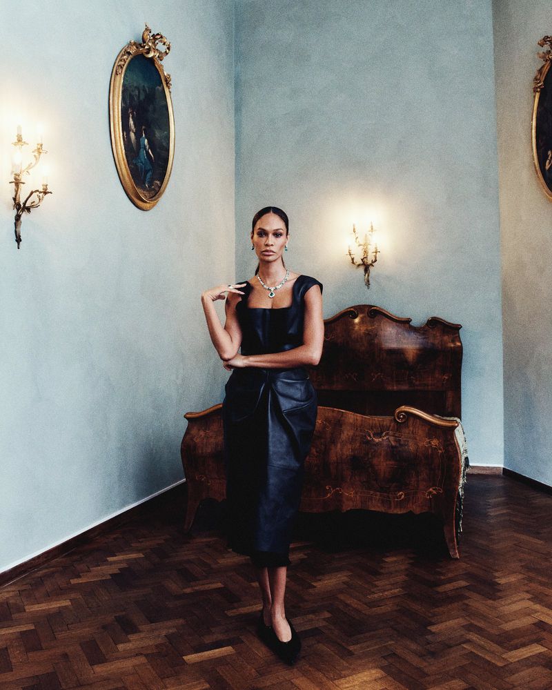 Adult woman in black dress and jewelry posing indoors against pale blue walls with classic paintings and wood parquet flooring.