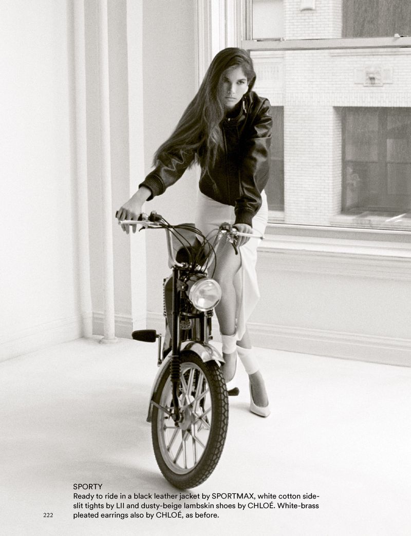 Woman in black leather jacket sits on a motorbike in a white photo studio with a brick wall visible through the window in the background.
