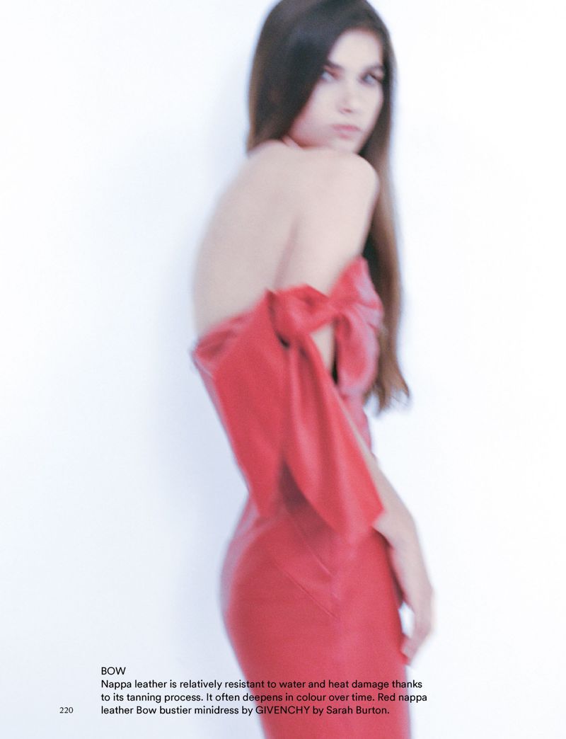 Brunette woman poses in a bright, studio shot wearing a red, off-the-shoulder leather dress with a bow detail and long, straight dark hair.
