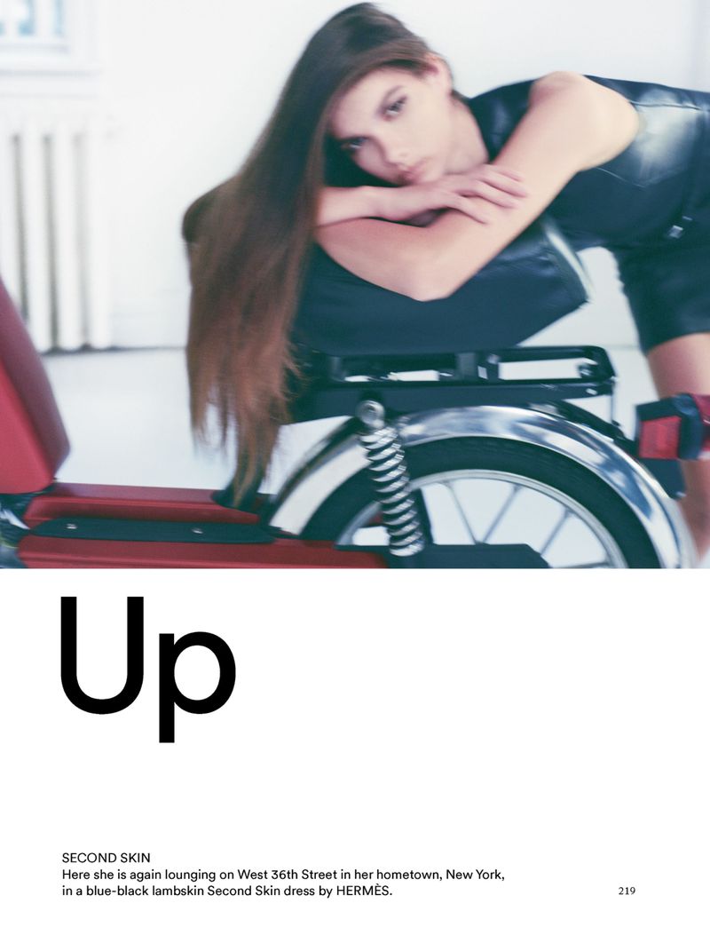 Brunette woman in black dress lounges on scooter with red accents in a photo studio. Image includes text "Up" and "Second Skin."