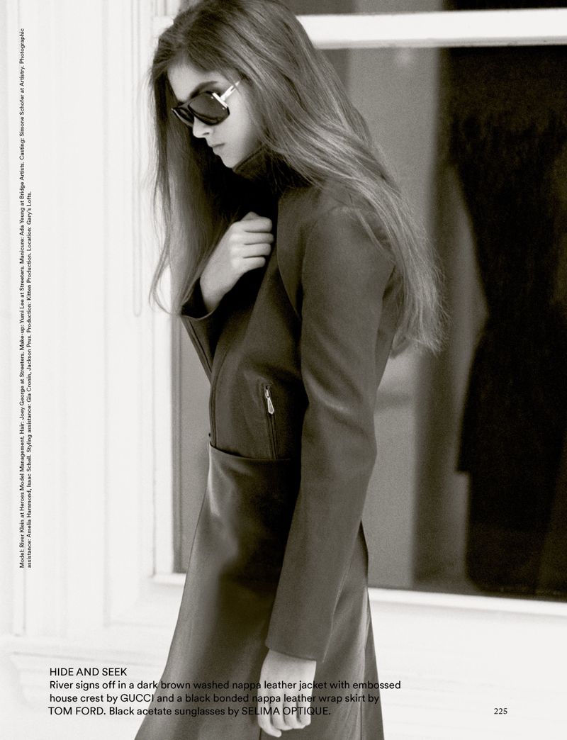 Fashionable woman in brown leather jacket and skirt, accessorized with sunglasses, poses in front of a window, exuding elegance.
