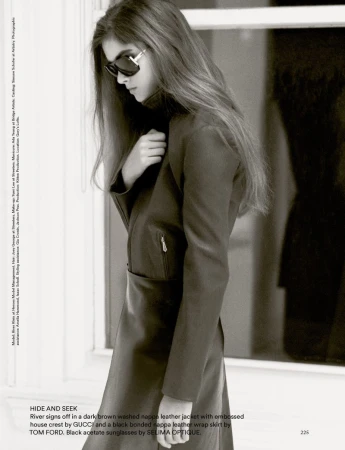 Fashionable woman in brown leather jacket and skirt, accessorized with sunglasses, poses in front of a window, exuding elegance.
