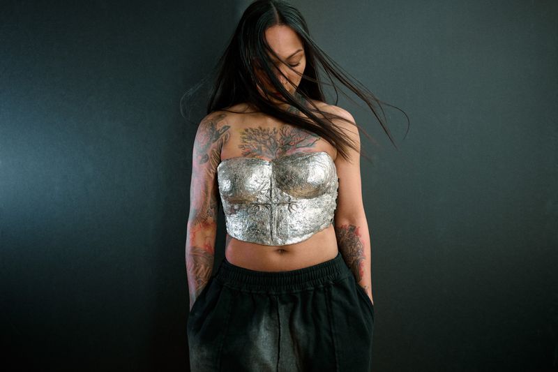 A young woman with a silver metallic top and black pants poses against a dark gray background, illuminated by soft studio lighting.