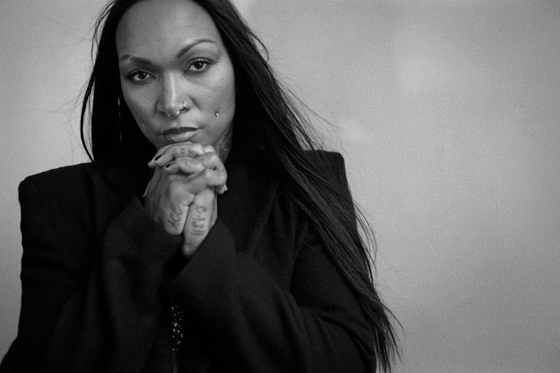 Black-and-white portrait of an adult female with dark hair and multiple facial piercings, wearing a dark coat and hands clasped, indoors.