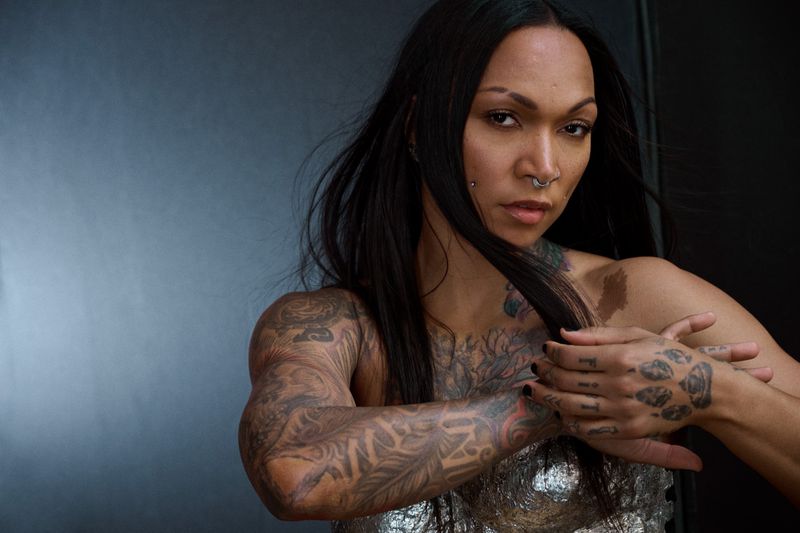 Portrait of adult woman with dark hair, tattoos, and nose ring, posing in a shiny, silver top against a dark gray background.