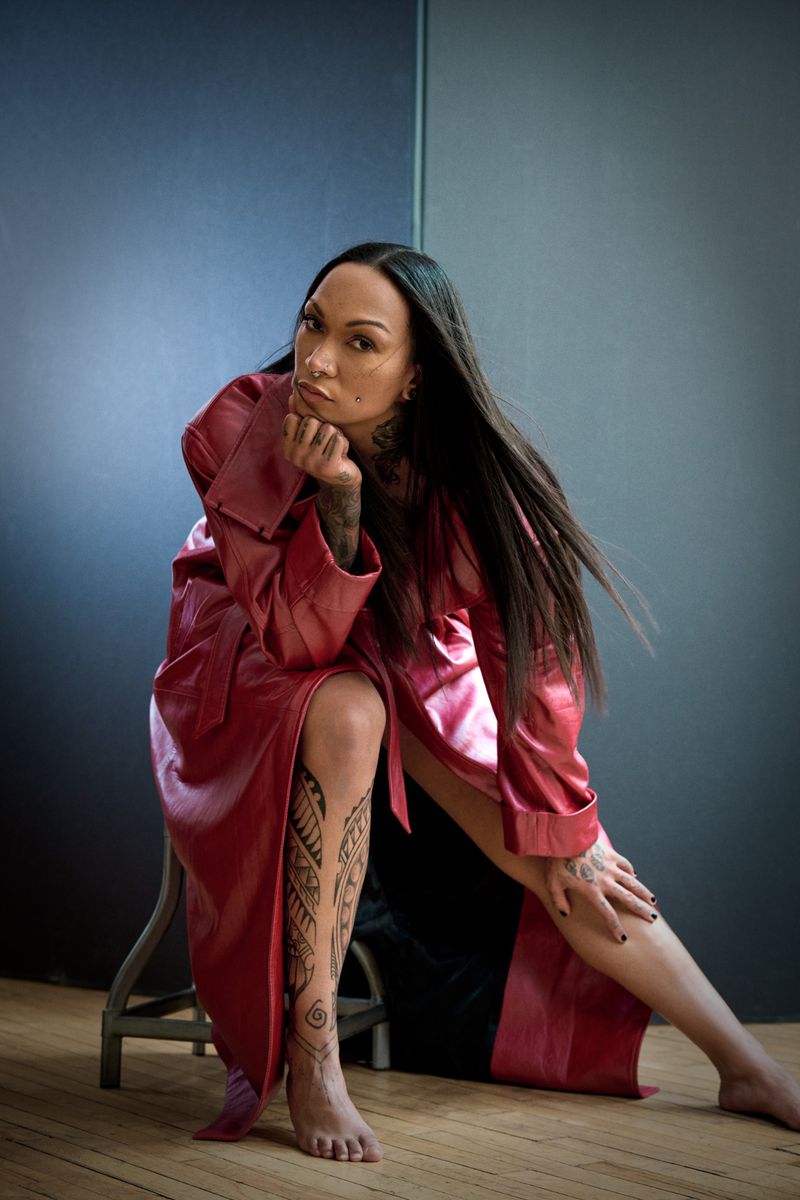 Woman with tattoos sits on stool wearing red coat. Tattoos on arms and leg are visible. Neutral background. Fashion shot.