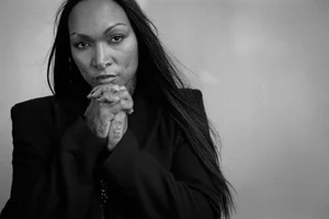 Monochrome portrait of a woman with long dark hair wearing a black jacket, hands clasped together near her face.