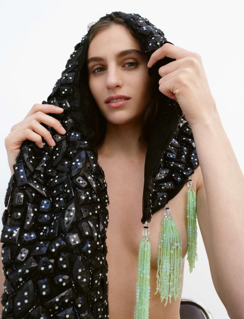A woman poses in a black, beaded hooded garment with flowing green tassels against a bright white backdrop in a studio setting.